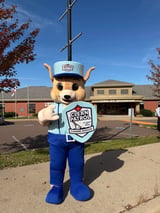 A large dog mascot wearing a blue sailor cap and Clean Patrol shirt stands on a sidewalk in front of a library building