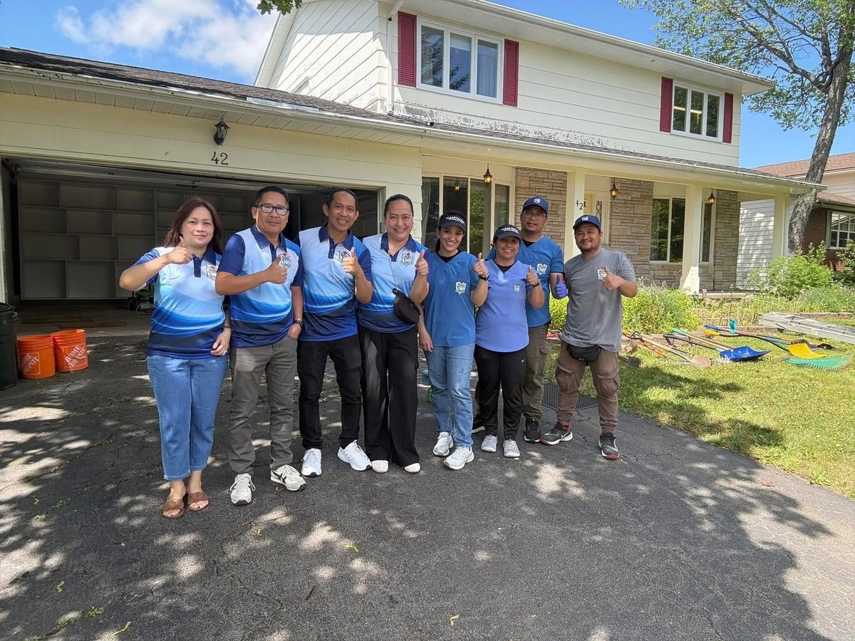 Group of volunteers in blue shirts posing together on a driveway in front of a house during a community service day