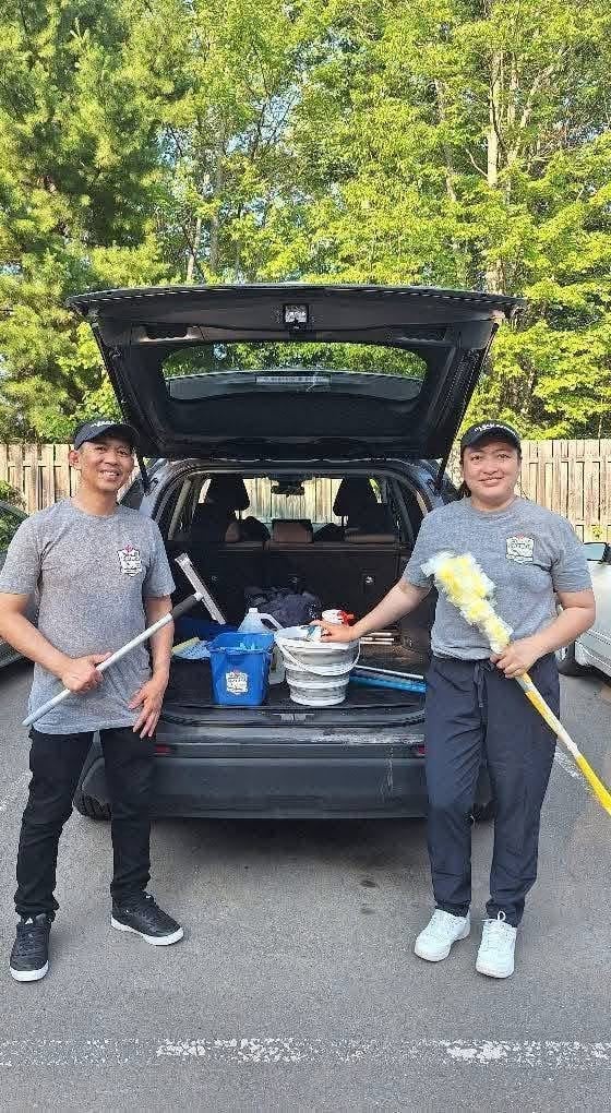 Two workers in matching t-shirts stand in front of an open car trunk filled with cleaning supplies, holding mops and a yellow duster
