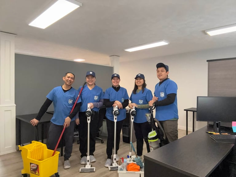 Five team members in blue shirts posing with cleaning equipment in an office break room